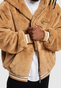 Kurtka bomberka z beżowego sztucznego futra z zamkiem błyskawicznym z przodu, prążkowanymi mankietami w paski oraz małym logo. Miękka w dotyku i o luźnym kroju.