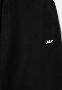 Czarna tkanina polarowa z białym wyhaftowanym napisem "Sports" w pobliżu dolnego, prawego rogu oraz widocznym bocznym szwem.