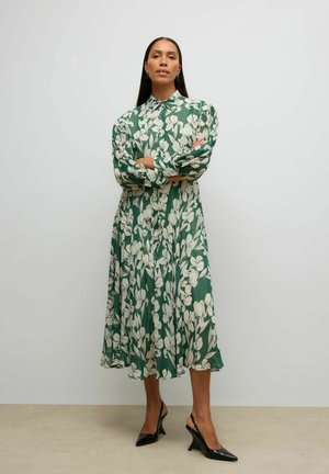 Nő áll karjait keresztbe téve, zöld midi ruhát visel fehér virágmintával és fekete pántos, alacsony sarkú cipőkkel.