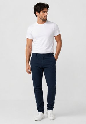 Mann trägt ein weißes T-Shirt, marineblaue Hose und weiße Turnschuhe, steht mit einer Hand in der Tasche und schaut nach links.