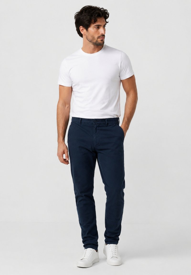 Man wearing a white t-shirt, navy blue pants, and white sneakers standing with one hand in pocket, looking to his left.