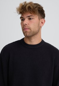 Jeune homme aux cheveux bouclés châtain clair et barbe naissante, portant un pull noir côtelé, regardant vers le bas et sur le côté sur un fond gris uni.