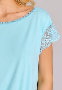Jasnoniebieska bluzka z krótkim rękawem, ozdobiona delikatnymi koronkowymi akcentami na rękawach oraz falbaniastym dekoltem, wykonana z miękkiego, gładkiego materiału.