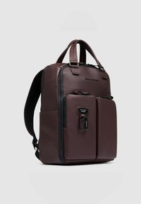 Zaino in pelle marrone con due tasche frontali, manici superiori e cinturini regolabili. Presenta cerniere e un dettaglio con targhetta in metallo.