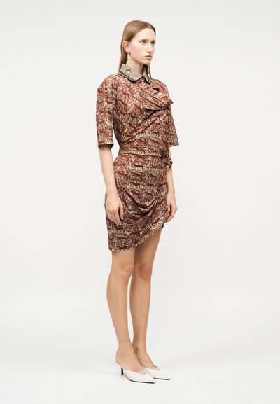 Γυναίκα που φοράει καφέ φόρεμα μέχρι το γόνατο με σχέδια και λεπτομέρειες ρυτιδώσεων, με αντίθετο γιακά, συνδυασμένο με λευκά παπούτσια χαμηλού τακουνιού.