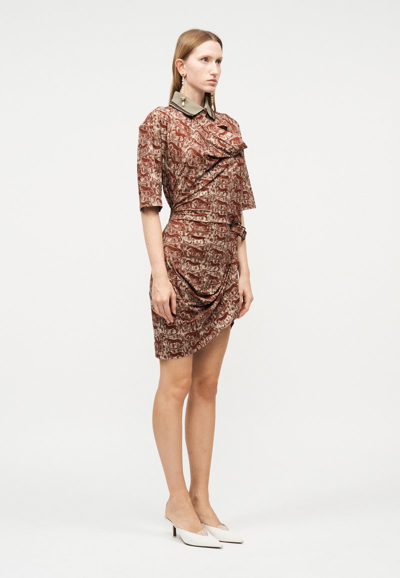 Donna che indossa un vestito marrone a fantasia di lunghezza fino al ginocchio con dettagli arricciati e un colletto a contrasto, abbinato a scarpe bianche con tacco basso.