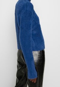 Pull bleu pelucheux à texture côtelée, coupe ajustée et manches longues, associé à un pantalon noir en fausse cuir brillant avec un motif crocodile.