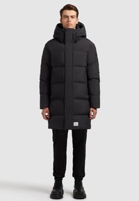 Hombre de pie con un abrigo largo de invierno acolchado negro con capucha, pantalones negros y botas negras, frente a un fondo claro y liso.