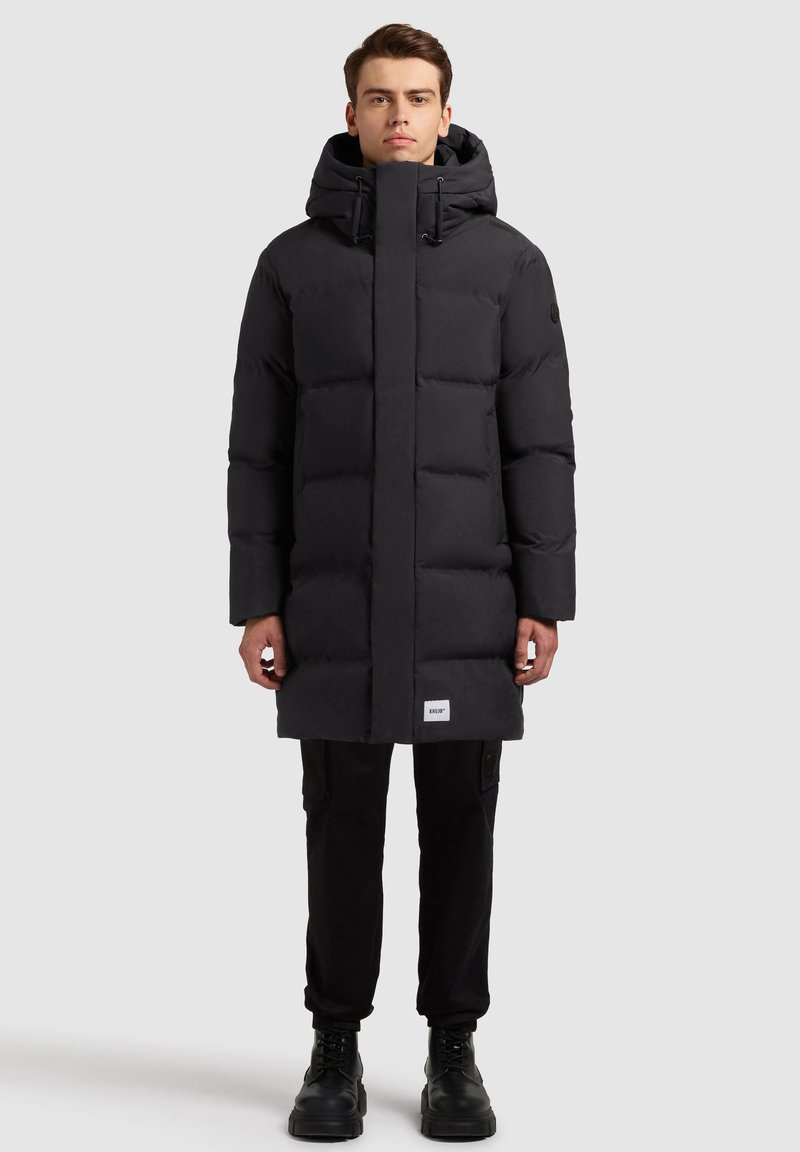 Hombre de pie con un abrigo largo de invierno acolchado negro con capucha, pantalones negros y botas negras, frente a un fondo claro y liso.