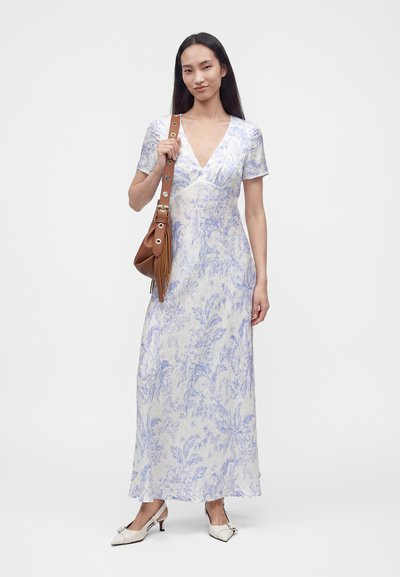 Femme portant une robe maxi fleurie blanche et bleu clair à manches courtes, des chaussures blanches à talons, et tenant un sac à main en cuir marron.