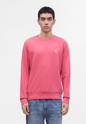Jeune homme portant un sweat-shirt uni rose à manches longues et un jean bleu, debout devant un fond blanc.