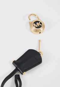 Lucchetto d'oro con un design circolare, caratterizzato da un centro nero e un logo inciso. Include un portachiavi in pelle nera e una chiave in metallo.