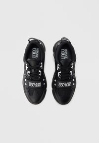 Černé sportovní tenisky mají texturovaný svrchní materiál z tkaniny, tkaničky a gumovou podrážku. Detaily zahrnují kontrastní bílé loga značky a nýty.