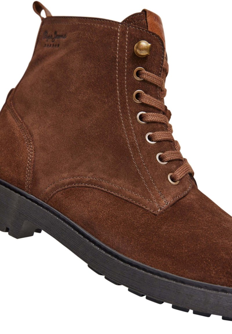 Botín de ante marrón con ojales de metal y cordones redondos. Presenta una suela de goma negra texturizada y un acento de cuero a contraste en el talón.