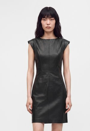Femme aux longs cheveux noirs portant une robe en cuir noir sans manches et moulante, debout devant un fond blanc uni.
