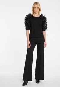 Schwarzes Oberteil mit gerafften, durchsichtigen Stoffärmeln, kombiniert mit eleganten schwarzen Hosen mit subtilen Verzierungen. Beide Kleidungsstücke sind figurbetont.