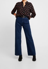 Camisa negra con estampado de animal rojo, cuello abotonado, combinada con jeans de pierna ancha de cintura alta en azul oscuro y botas negras con tobillo.