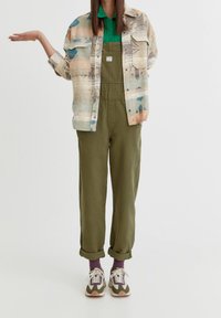 Chemise multicolore à boutons avec motifs abstraits, chemise verte à col, salopette olive et revers de manches retroussés. Associée à des baskets et des chaussettes violettes.