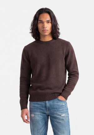 Giovane uomo con capelli neri fino alle spalle che indossa un maglione marrone e jeans blu strappati, in piedi con una mano in tasca su sfondo bianco.