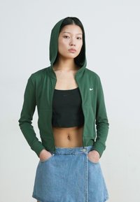 Zelená mikina na zips vyrobená z mäkkého materiálu, má kapucňu a bočné vrecká. Nosená cez čierny krátky top a džínsovú sukňu.