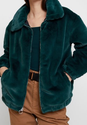 Femme portant une veste en fausse fourrure vert foncé avec fermeture éclair, un haut côtelé couleur sarcelle et un pantalon marron avec une ceinture noire, mains dans les poches de la veste.
