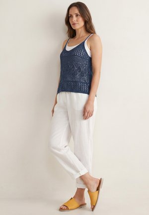 Frau trägt weiße Hose, weißes Tanktop, dunkelblaues gehäkeltes ärmelloses Oberteil und gelbe Slip-on-Sandalen und steht an einer schlichten Wand.