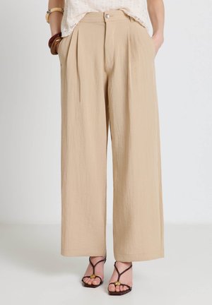 Femme portant un pantalon beige à jambes larges avec des plis et une fermeture à boutons, assorti à des sandales à brides foncées et un haut en dentelle blanche.
