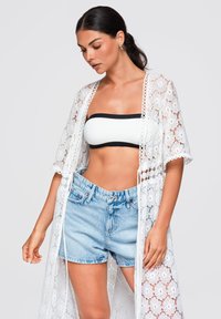 Femme portant un cardigan blanc en dentelle ouvert, un haut bandeau blanc avec bordure noire et un short en jean bleu clair sur un fond uni.