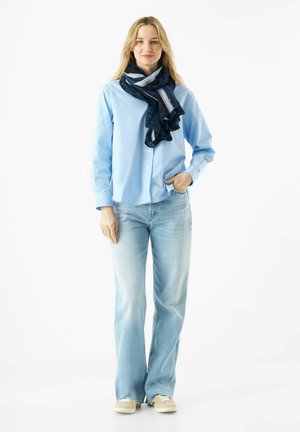 Lichtblauw overhemd met knopen, gecombineerd met losse, lichtblauwe jeans en een donker patroonde sjaal met strepen in blauw en pink.