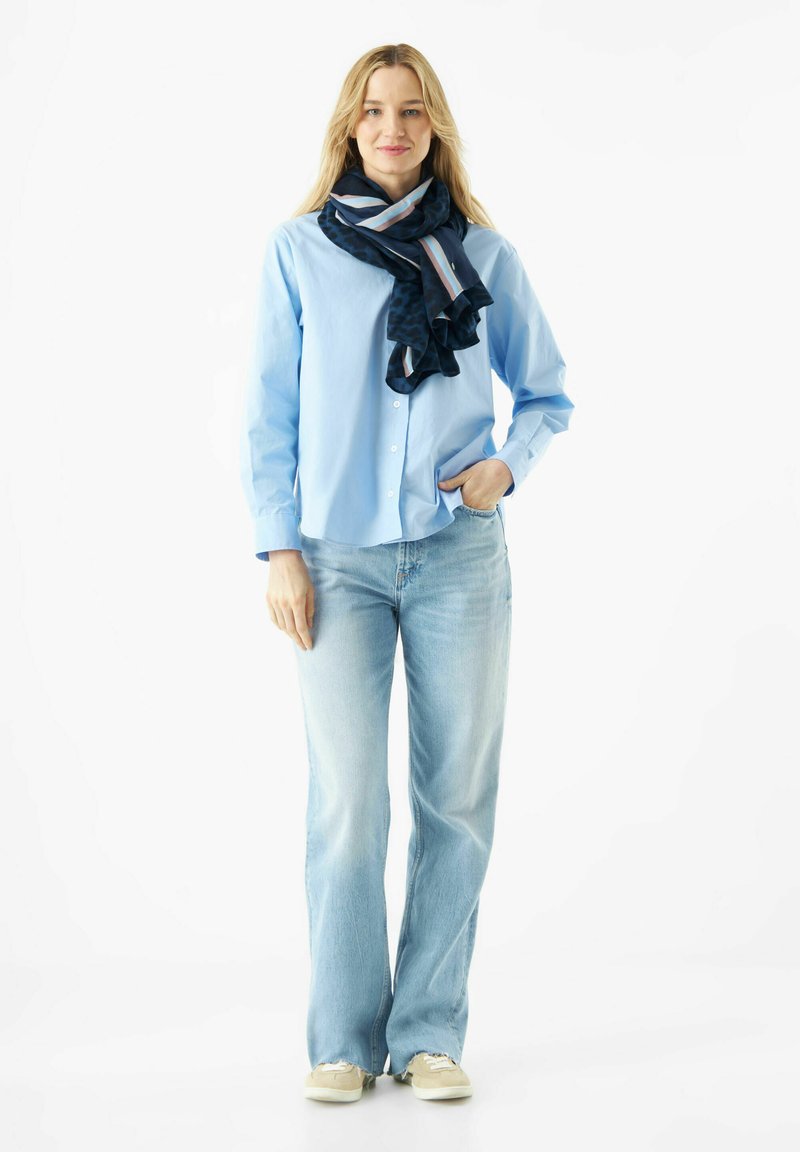 Chemise à boutons bleu clair associée à un jean ample bleu clair et une écharpe à motifs sombres avec des rayures bleues et roses.