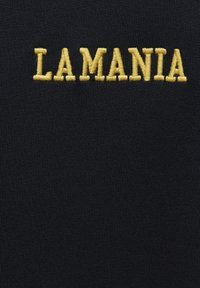 Čierna tkanina s výrazným žltým vyšívaným textom, ktorý znie "LA MANIA." Textúra je hladká s mierne rebrovaným vzhľadom.