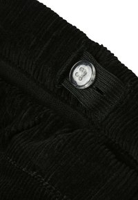 Le tissu en velours côtelé noir présente des côtes verticales avec un bouton transparent et un détail de taille cousu. La vue de près met en évidence la texture.