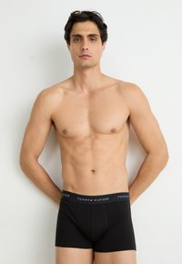 Fekete Tommy Hilfiger boxer alsónadrág, feliratozott elasztikus derékpánttal. A modellen rövid sötét haja van és semleges kifejezésű, egy sima háttér előtt áll.