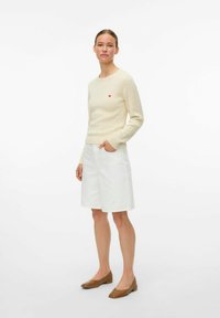 Romig geribde gebreide trui met een klein rood hartlogo, gecombineerd met witte denim short en bruine puntige flats. Eenvoudige, casual outfit.