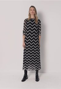 Frau trägt ein langärmliges Midikleid mit schwarz-weißem Zickzackmuster, kombiniert mit schwarzen Stiefeletten, steht vor einem schlichten Hintergrund.