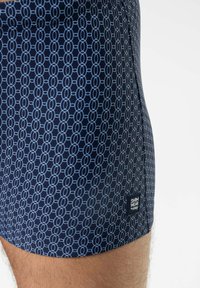Marineblaue Badeshorts mit einem geometrischen hellblauen Muster aus ineinandergreifenden Kreisen. Versehen mit einem kleinen schwarzen Etikett mit der Aufschrift "SWIM WEAR by mey."