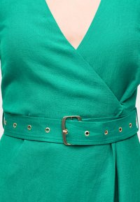 Πράσινο φόρεμα τύπου wrap με V-λαιμόκοψη και φαρδιά ζώνη που διαθέτει χρυσή αγκράφα και λεπτομέρειες με μεταλλικούς κρίκους γύρω από τη μέση.