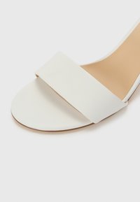 Hvit sandaler med bred rem over tærne, flat såle og glatt lærtekstur. Enkel design, åpen hæl og minimale detaljer.
