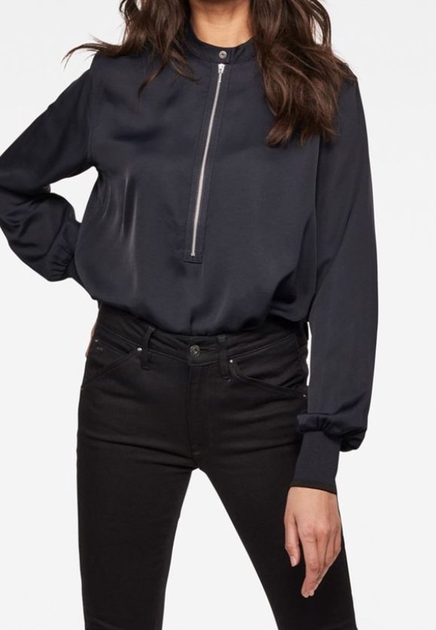 Femme portant un chemisier noir à manches longues avec une fermeture éclair à l'avant et un col montant, associé à un pantalon noir ajusté.