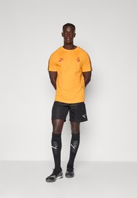 Oranje sportshirt met korte mouwen met de tekst "KING" en logo; zwarte shorts; zwarte scheenbeschermers; zwarte sportschoenen met witte details.