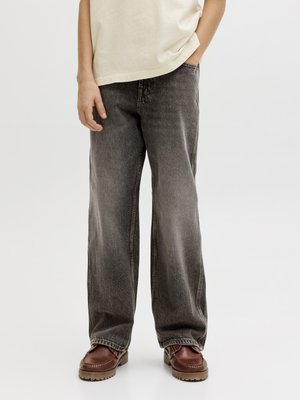 Oseba, ki nosi široke sive oprane kavbojke, rjave usnjene čevlje in kremno barvno srajco, z eno roko v žepu.