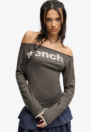 Ung kvinde med flettet mørkt hår bærer en off-shoulder langærmet top med "Bench" logo og lagdelt navy nederdel mod hvid baggrund.