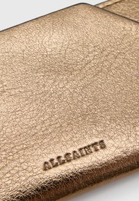 Cartera de cuero metálico dorado con acabado texturizado, que presenta un logo en relieve de "ALLSAINTS". Los bordes son suaves, con un diseño minimalista.
