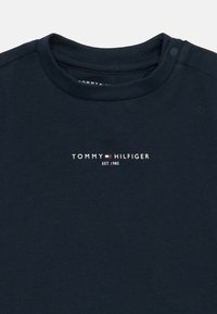 T-shirt de algodão azul marinho com decote redondo e botão de pressão. A parte frontal exibe "TOMMY HILFIGER EST. 1985" em texto branco.