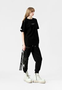 Camiseta negra de gran tamaño con un bolsillo en el frente y detalle de raya lateral, combinada con pantalones jogger negros con un patrón de hojas texturizado y bolsillos. Botas de combate blancas.