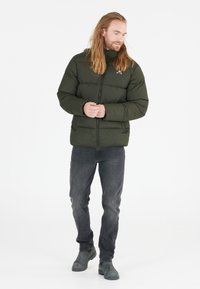 Puffjakke i mørkegrøn, med lynlås-lukning, ribbede ærmer og et diskret logo. Sætter sammen med grå jeans og grå støvler.