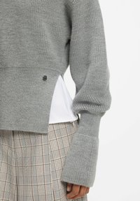 Close-up of a person wearing a grey knit sweater with flared cuffs, a white shirt underneath, and beige plaid pants.