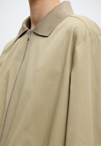 Inimese lähedalt võetud foto, kes kannab beeži värvi lukuga jopet soonikkoelise kraega ja langetatud õlasaumadega.