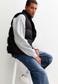 Czarna kamizelka puchowa z wysokim kołnierzem, noszona na jasnoszarym longsleeve'ie. Ciemnoniebieskie dżinsy i białe sneakersy dopełniają strój.