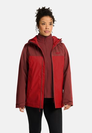 Frau mit lockigem Haar, die eine rot-bordeauxfarbene Reißverschlussjacke und schwarze Hose trägt, steht vor einem einfarbigen hellen Hintergrund.